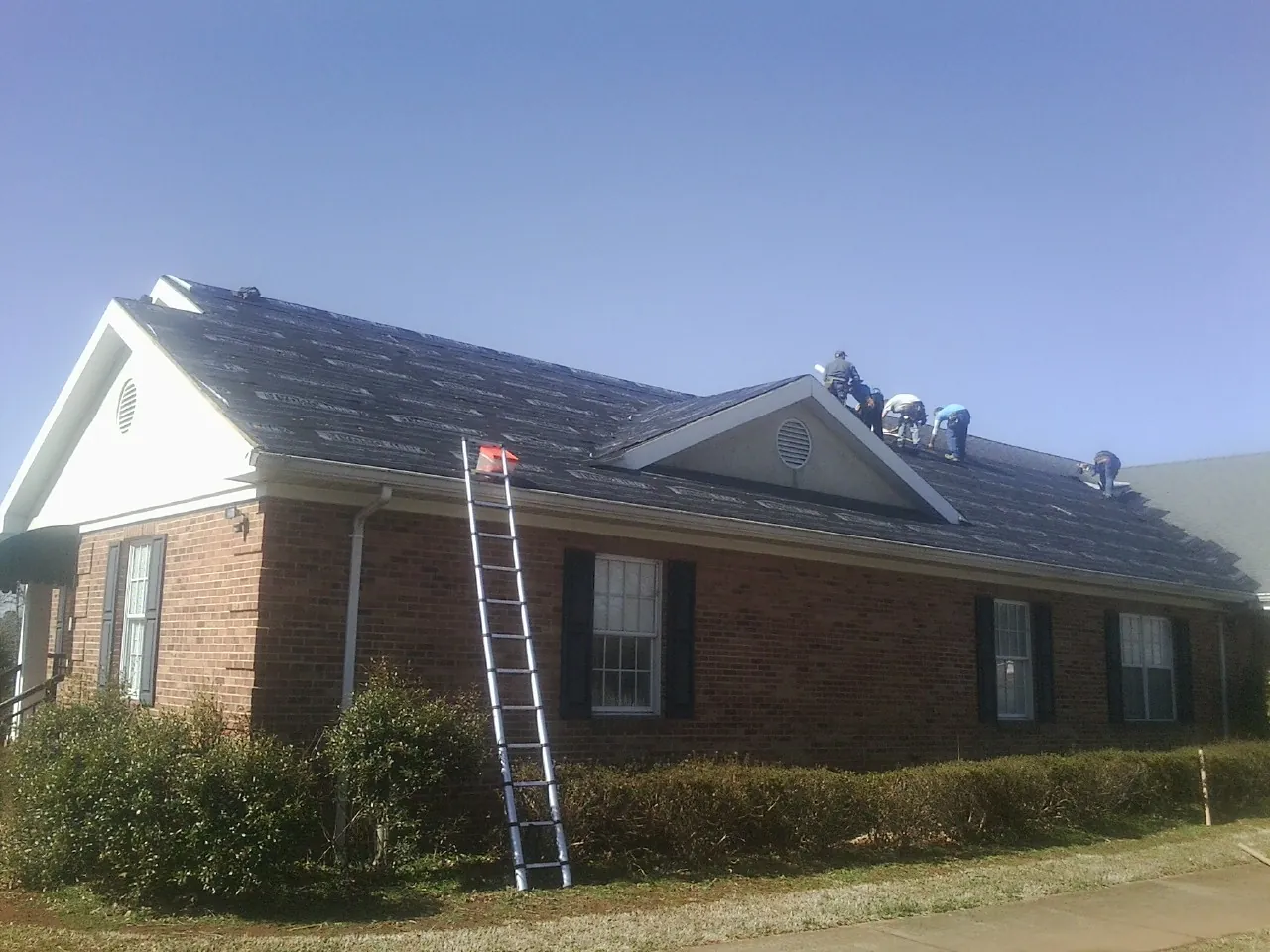 Crew tearing shingles off a brick ranch roof with a ladder set — Emergency Roof Repair work by our crew in Conroe
