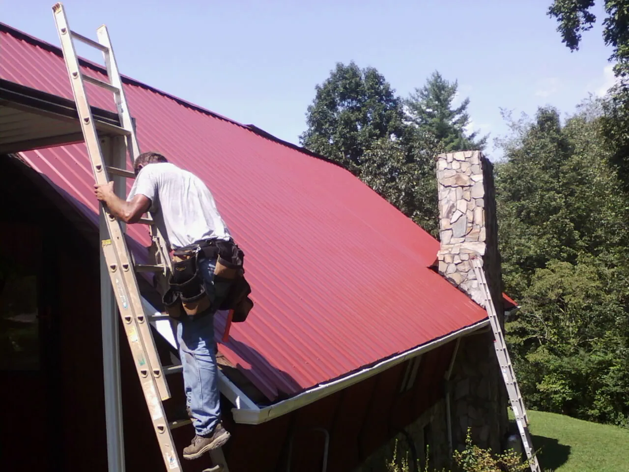 Roofer on a ladder beside a red metal roof and stone chimney — Soffit & Fascia Repair by Roofing Brothers in Conroe